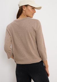 Femme portant une chemise beige à manches longues, un pantalon noir et une casquette de baseball crème, se tenant devant un fond blanc uni.