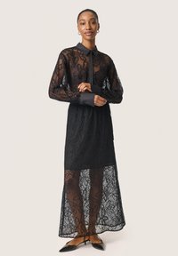 Abito lungo nero in pizzo con motivo floreale, colletto, chiusura con bottoni e sovrapposizione di gonna trasparente. Tacchi a punta neri completano il look.