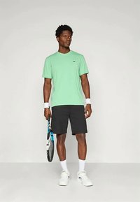 Zelené tričko s krátkym rukávom, čierne šortky, biele ponožky a tenisové topánky. Drží tenisovú raketu s modrými akcentmi. Jednoduchý dizajn, voľný strih.