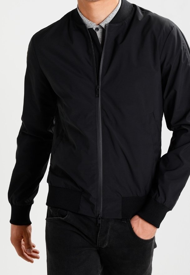 Homme portant une veste bomber noire zippée par-dessus une chemise grise à boutons, une main dans la poche et l'autre détendue le long du corps.