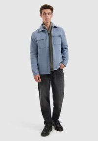 Lichtblauwe wollen jas met twee borstzakken, knoopsluiting en kraag; gelaagd over een grijze vest met rits; gedragen met donkere jeans en zwarte schoenen.