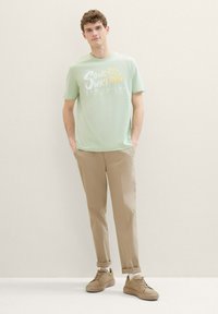 Ljusgrön t-shirt med kort ärm och texten "Soul Surfing", tillsammans med beige byxor och bruna sneakers, presenterad på en neutral bakgrund.