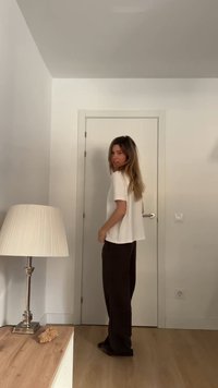 Femeie îmbrăcată într-o cămașă albă și pantaloni închiși la culoare, stând lângă o ușă închisă într-o cameră minimalist decorată, cu o lampă pe o masă din lemn.