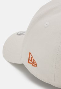 Hellbeige Baseballkappe mit gebogenem Schirm, strukturiertem Stoff und einem bestickten orangefarbenen Logo an der Seite. Die Nähte sind entlang der Ränder sichtbar.