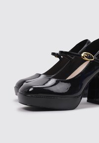 Zapatos de tacón Mary Jane de charol negro con punta redonda, plataforma gruesa y tacón bloque robusto; presentan una hebilla en tono dorado en la correa.