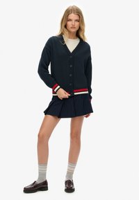 Marineblauwe cardigan met knoopsluiting, geribde kraag en manchetten met rode en crème strepen. Gecombineerd met een geplooide krijtstreeprok en loafers.