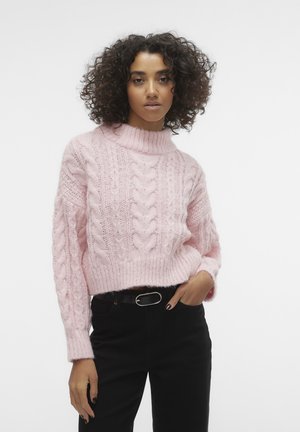 Rosa stickad tröja med hög krage, med ett kabelstickat mönster och ribbat avslut. Kombinerad med svarta byxor och ett svart bälte.