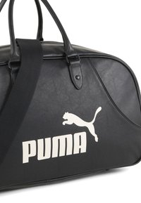Sac de sport en simili cuir noir avec des poignées solides et une sangle réglable, arborant un logo Puma blanc et un design minimaliste.