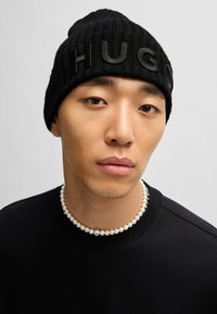 Svart strikket lue med hevet tekst "HUGO" på forsiden; brukt sammen med en enkel svart skjorte og en perlehalsskjede.