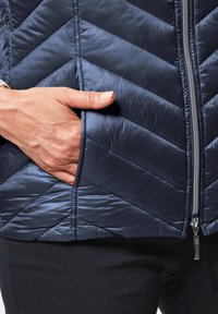 Marineblaues, isoliertes Weste mit Chevron-Muster, strukturiertem, glänzendem Stoff, Reißverschluss vorne und einer Hand in der Seitentasche.