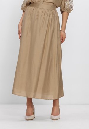 Vrouw draagt een beige rok tot de enkel met een hoge tailleband, gecombineerd met beige hoge hakken, staand tegen een effen achtergrond.
