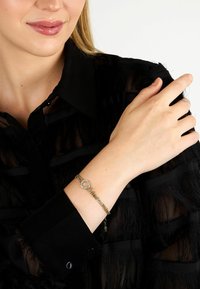 Bracelet chaîne en or avec un fermoir circulaire orné de petites pierres claires, porté sur un poignet avec un chemisier noir texturé.