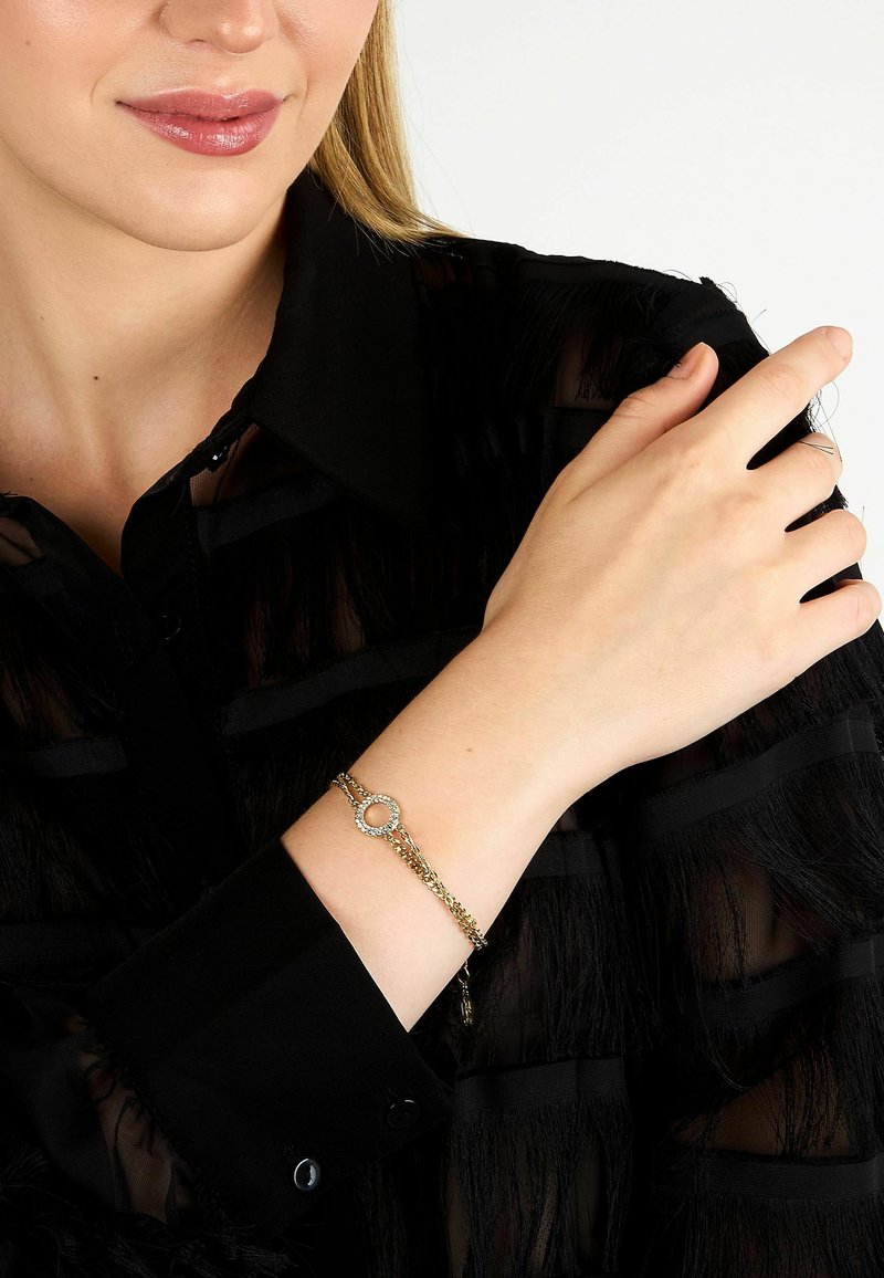 Bracelet chaîne en or avec un fermoir circulaire orné de petites pierres claires, porté sur un poignet avec un chemisier noir texturé.