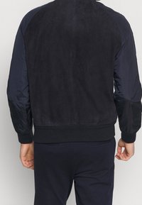 La cazadora bomber azul marino presenta un respaldo de ante, mangas de tela suave, puños y dobladillo acanalados. Diseño sencillo con un ajuste relajado.
