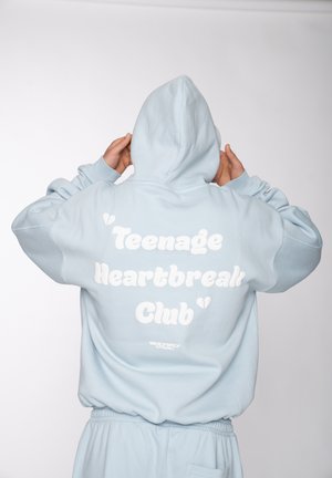 Person trägt hellblauen Hoodie und Hose, hält die Kapuze, mit dem Text „Teenage Heartbreak Club“ auf dem Rücken vor einfarbigem Hintergrund.