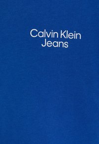 Modrá bavlněná t-shirt s logem "Calvin Klein Jeans" v bílé barvě, umístěným uprostřed v moderním písmu, s hladkou texturou a klasickým střihem.