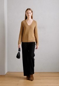 Pull en tricot beige à col en V, associé à un pantalon large noir. Le mannequin tient un petit sac à main noir et porte des bottines marron.