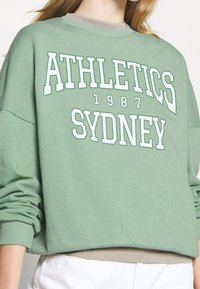 Sudadera verde de algodón con la inscripción "ATHLETICS SYDNEY 1987" en blanco, de diseño oversize, puños acanalados y cuello redondo.
