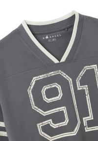 Camiseta gris de manga corta con escote en V y ribete color crema. Presenta grandes números blancos "91" desgastados y dos rayas blancas. Tejido ligero.