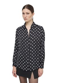 Camicia nera in tessuto leggero con pois bianchi, maniche lunghe, chiusura frontale con bottoni e orlo leggermente curvo. Indossata con una gonna nera.