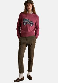 Naine kannab pruuni ruudulisi pükse, valgeid sokke, pruune lohvakleidimude ja roosa naistekirjutusega sviitrit, millel on lilleline jeepi muster, ning pruuni mustrilist kübarat.