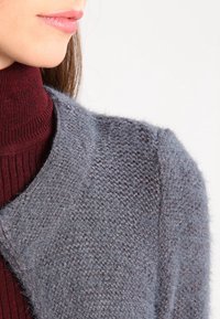 Žena v burgundském vroubkovaném roláku pod texturovaným šedým kardiganem, oříznutá tak, aby byly vidět rty a rameno.