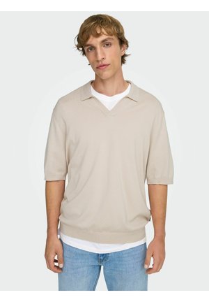 Giovane uomo con capelli castano chiaro che indossa un maglione beige a maniche corte sopra una camicia bianca e jeans blu, in piedi davanti a uno sfondo bianco.