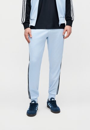 Mand iført lyseblå træningsbukser med sorte sidelister, mørkeblå Adidas-sneakers og en sort og lyseblå jakke med hvide striber.