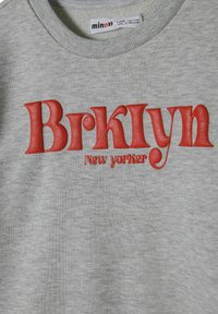 Szürke pulóver kiemelkedő piros betűzéssel, amelyen ez áll: "Brklyn New Yorker." A szövet lágy tapintású, bordázott nyakkal és mandzsettával rendelkezik.