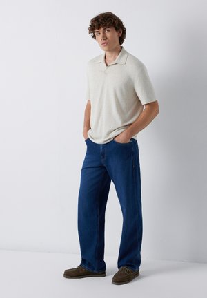 Young man standing with hands in pockets wearing a light beige short-sleeve polo, blue jeans, and brown lace-up shoes against a white background.