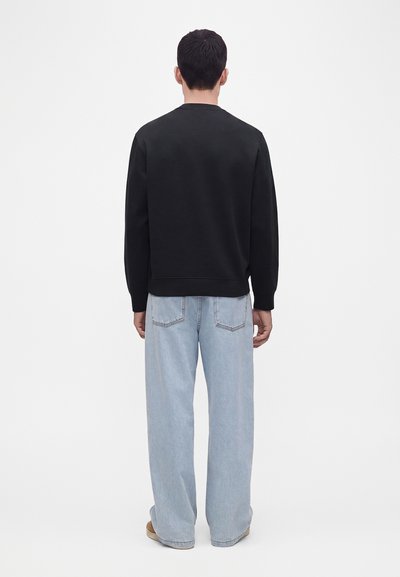 Homme debout tourné de dos, portant une chemise noire manches longues et un jean bleu clair à jambes larges sur un fond uni.
