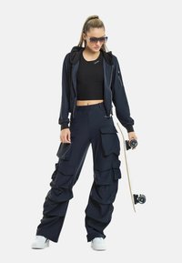 Marina cargo-byxor med flera fickor och en avslappnad passform, i kombination med en åtsittande svart kort topp och en marinblå zip-up-jacka; modellen håller en skateboard.