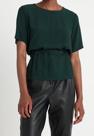 Ciemnozielona bluzka z krótkim rękawem ze ściąganym w talii dołem, zestawiona z czarnymi, wysokim stanem skórzanymi spodniami na stojącej osobie.