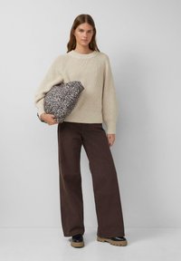 Frau trägt einen beigen Strickpullover, weite braune Hose und schwarze Stiefel, während sie eine Tasche mit Leopardenmuster vor einem schlichten grauen Hintergrund hält.