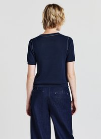 Marineblaues, gestricktes Oberteil mit kurzen Ärmeln und rundem Ausschnitt, verziert mit hellen Steppnähten. Kombiniert mit Jeanshosen.