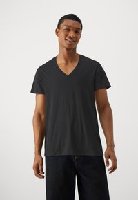 Tricou din bumbac negru, cu un gât în formă de V, mâneci scurte și o croială lejeră. Prezintă un branding minim pe piept. Textură netedă.