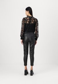 Blusa negra transparente con bordados florales y lentejuelas, combinada con pantalones de imitación cuero negros y zapatos de tacón grueso. Vista trasera.