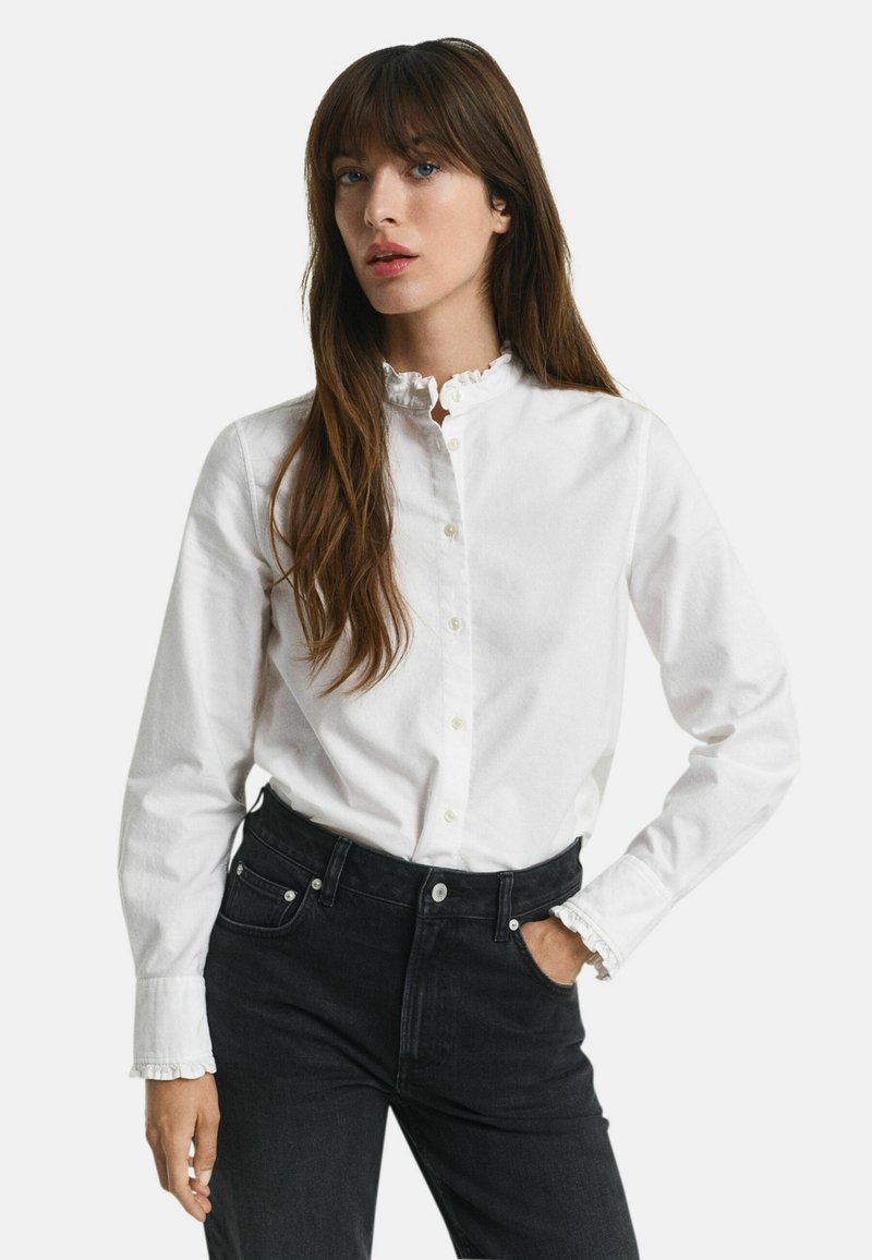 Camicia bianca con colletto e polsini arricciati, maniche lunghe e vestibilità classica, abbinata a jeans neri a vita alta.