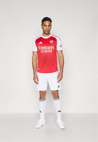 Rode en witte voetbaltrui met "Emirates Fly Better" gedrukt op de voorkant, gecombineerd met witte shorts en sokken met zwarte strepen.