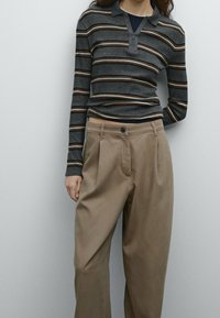 Szara w paski koszulka polo z długim rękawem i zamkiem przy kołnierzu, zestawiona z beżowymi spodniami typu wide-leg z przodu z plisami i zapięciem na guzik.