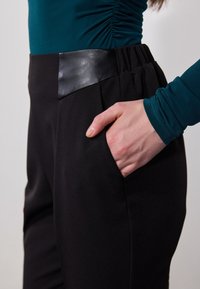Black pants with a smooth leather waistband and a pocket, paired with a teal long-sleeve top featuring gathered detailing at the shoulder.