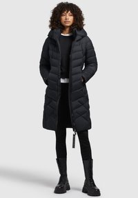 Manteau en duvet noir avec une capuche, présentant une couture en forme de losange, une fermeture éclair intégrale et des poches latérales. Porté sur un haut noir et un pantalon noir.