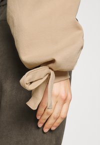 Manche longue beige avec un nœud noué au poignet sur le bras d'une personne appuyée contre un pantalon foncé.