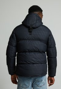 Chaqueta acolchada negra con un patrón geométrico, que incluye capucha y correa ajustable en la parte posterior. Combinada con jeans desgastados de color azul claro.