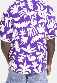 Man draagt een paarse shirt met korte mouwen met grote witte abstracte plant- en bladvormen, gecombineerd met lichtblauwe denim shorts.