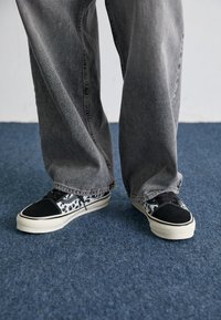 Mustavalkoiset kuvioidut tennarit, joissa on sileä musta kärkiosa, valkoinen pohja ja teksturoidut nauhat, yhdistettynä väljiin harmaisiin farkkuihin.