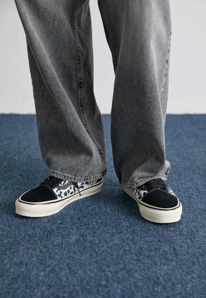Baskets à motifs noir et blanc avec un bout lisse noir, une semelle blanche et des lacets texturés, associées à un jean gris ample.