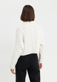 Pull en maille blanche avec un col haut, une coupe courte et un ourlet côtelé. Porté avec un pantalon noir taille haute.