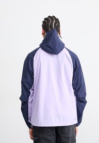 Veste coupe-vent légère violette et marine avec capuche, présentant un design de blocs de couleur et des poignets élastiques, vue de dos.