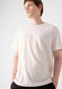 Ljusbeige t-shirt med rund halsringning, tillverkad av mjukt, strukturerat tyg med korta ärmar; har en subtil logotyp på bröstet.